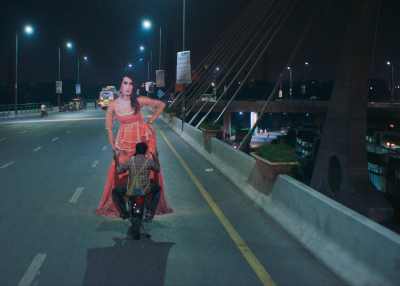 A man riding a motorcycle down a highway at night while carry a very large (twice his size) cardboard cutout of a dancer.